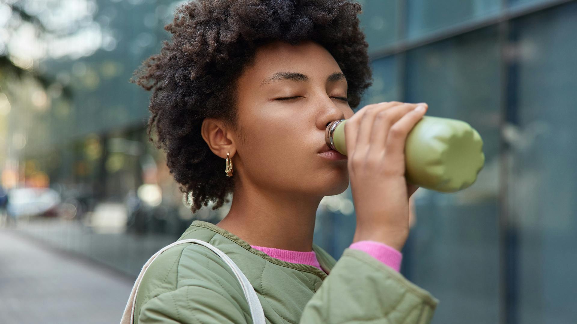 Tired thirsty woman with Afro hair drinks water from bottle takes break while walking in city poses in urban setting keeps eyes closed wears jacket hydrates herself. People lifestyle wellbeing concept